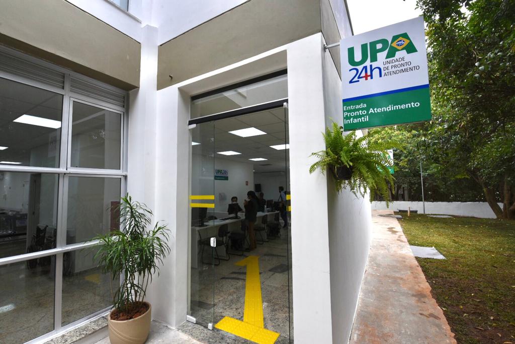 A entrada do prédio da ala infantil da UPA tem portas de vidro transparente de onde se vê uma mesa com pessoas na recepção, A porta da direita está aberta e tem um vaso de plantas à sua esquerda